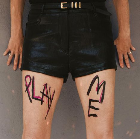 Auf den Oberschenkeln steht "PLAY ME" in schwarzer und pinker Schrift. Eine Person trägt schwarze Shorts.