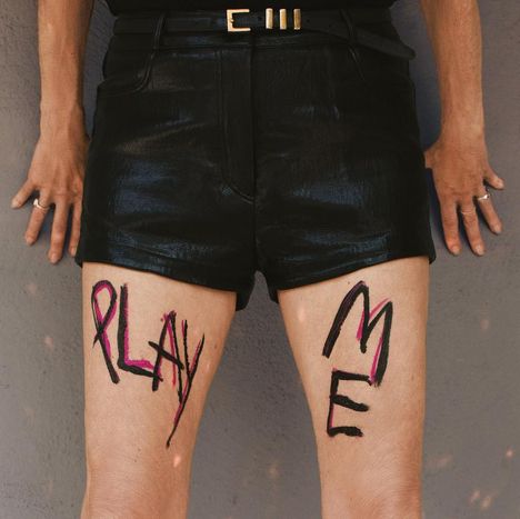 Auf den Oberschenkeln steht "PLAY ME". Eine Person trägt schwarze Ledershorts und hat Ringe an den Fingern.