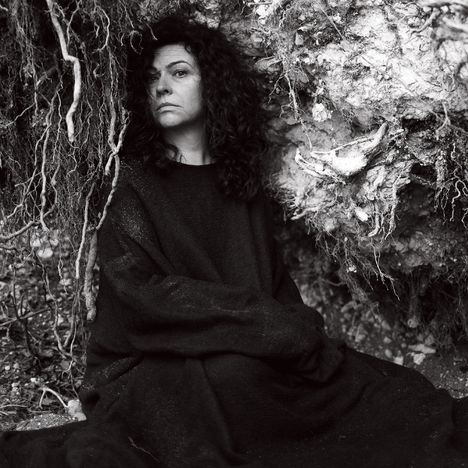 Es gibt keinen Text. Eine Person mit lockigem Haar sitzt in einem dunklen Kleid neben einem umgestürzten Baum mit freiliegenden Wurzeln.