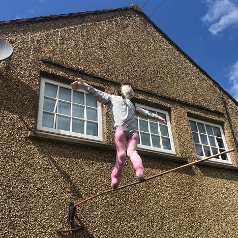 Eine Schaufensterpuppe in rosa Leggings und grauem Oberteil balanciert auf einem Seil vor einem Haus mit brauner Wand und Fenstern.