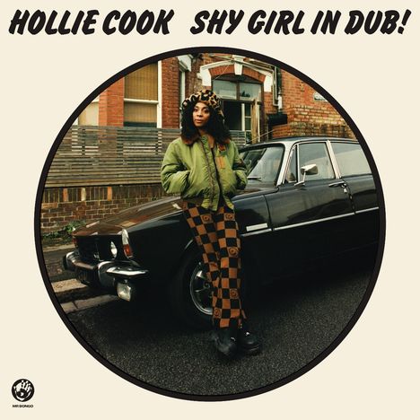 „HOLLIE COOK SHY GIRL IN DUB!“ Frau in grüner Jacke und karierter Hose lehnt an einem schwarzen Auto auf einer Straße.