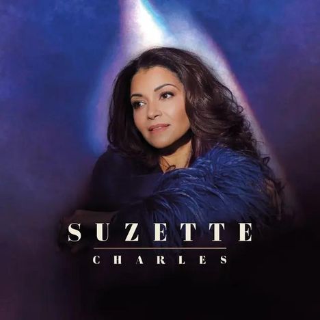 Text: "SUZETTE CHARLES". Eine Frau mit langen, lockigen Haaren und einem blauen Federumhang vor einem dunkelblauen Hintergrund.