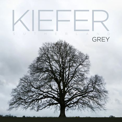 "Kiefer Sutherland, GREY" in großen Buchstaben. Ein kahler Baum unter grauem Himmel, ruhig und imposant.