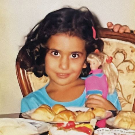 Ein Kind mit lockigem Haar hält eine Puppe. Vor ihm liegt Gebäck auf einem Tisch. Es lächelt leicht.