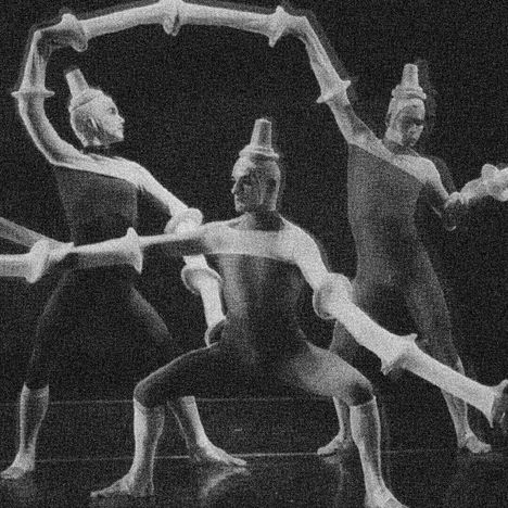 Drei Personen in Kostümen mit abstrakten, röhrenförmigen Strukturen, die eine Skulptur oder Skizze nachahmen.