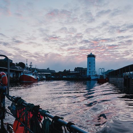Ein ruhiger Hafen bei Sonnenuntergang, rote Boote, ein weißer Turm, und reflektierendes Wasser mit wolkigem Himmel.