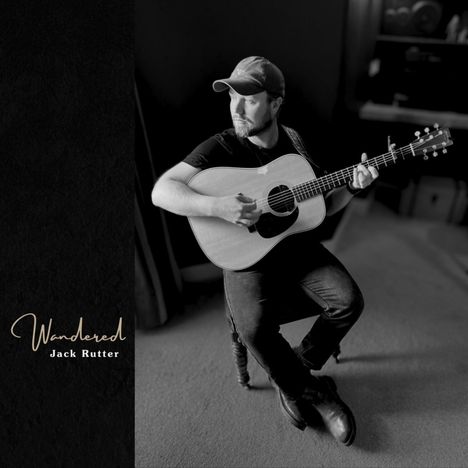 "Wandered. Jack Rutter." Ein Mann sitzt mit Gitarre in einem dunklen Raum, trägt eine Mütze und schaut nach rechts.