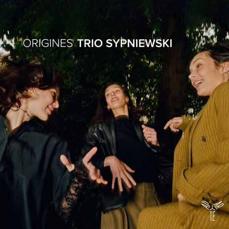 ORIGINES TRIO SYPNIEWSKI. Drei fröhliche Frauen tanzen in einem Wald. Unten rechts kleines weißes Logo einer Trompete.