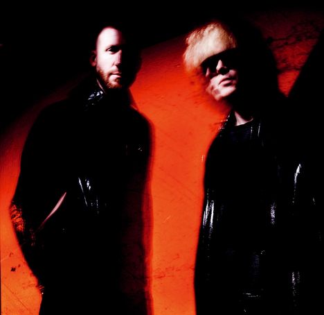 Zwei Personen vor einem intensiven roten Hintergrund, gekleidet in dunkle Kleidung, eine Person trägt eine Sonnenbrille.