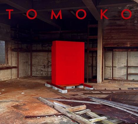 "TOMOKO" in großen roten Buchstaben. Im Hintergrund ein leerer, verlassener Raum mit einem roten Quader in der Mitte.