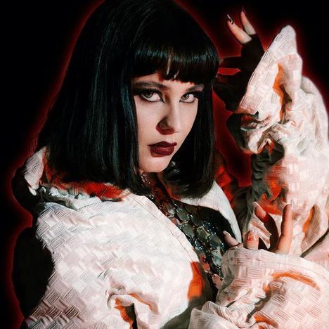 Eine Frau mit schwarzem Haar und dramatischem Make-up, in einem strukturierten weißen Mantel, vor einem rot beleuchteten Hintergrund.