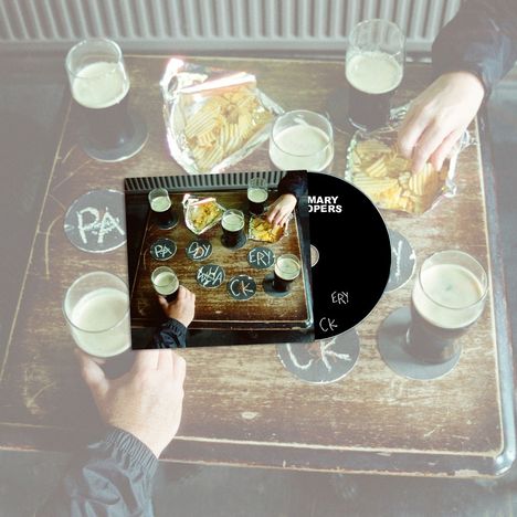 Kreise auf Tisch: "PA", "DY", "ERY", "CK". Hände mit Bier, Schalen mit Chips auf Holztisch. CD mit Teil der Szene.