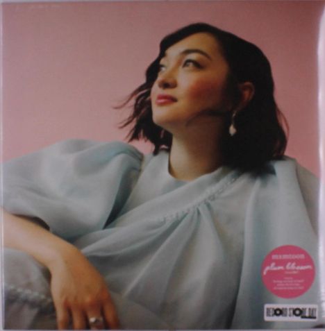 Aufschrift auf rundem, rosa Sticker: "AMMONS plum blossom." Frau in blassblauem Kleid vor rosa Hintergrund.