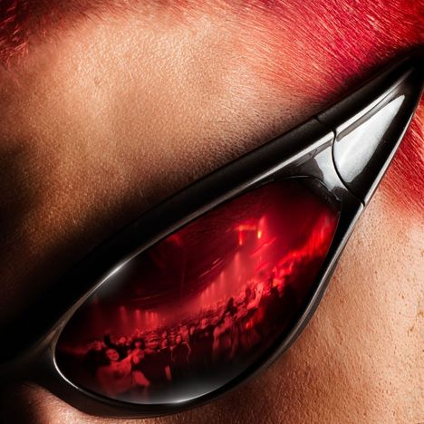 Es gibt keinen sichtbaren Text. Nahaufnahme eines Gesichts mit rotem Haar, spitze Sonnenbrille mit Spiegelung einer Menschenmenge.