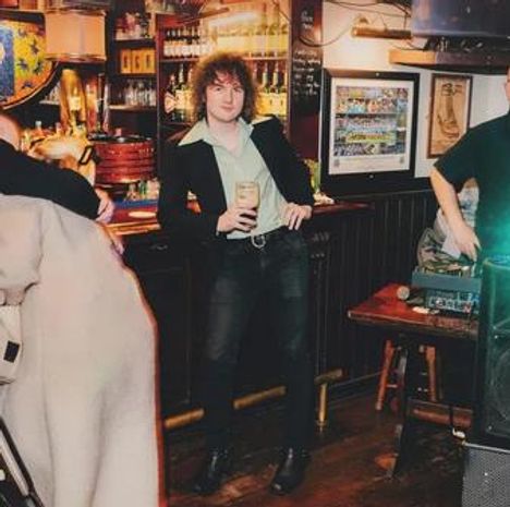 Ich kann die Texte nicht lesen. Eine Person steht lässig in einer gemütlichen Bar und hält ein Getränk in der Hand.