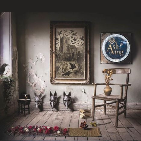 „Ash Wing“ in einem runden Logo. Alter Raum, Gemälde an der Wand, Holzstuhl, Tiermasken, Bücher, Blumen, ein Rabe am Fenster.