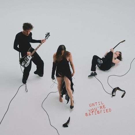 „UNTIL YOU'RE SATISFIED“ in roter Schrift. Drei Musiker: Bassist, Sängerin in Schwarz und Gitarrist auf dem Boden.
