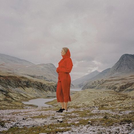 Kein Text vorhanden. Eine Person in rotem Outfit steht in einer bergigen Landschaft mit einem Fluss im Hintergrund.