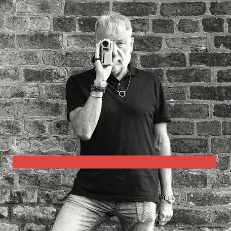 Roter Balken, schwarz-weißes Foto. Mann mit Kamera vor Gesicht, trägt ein dunkles Shirt, steht vor Ziegelmauer.