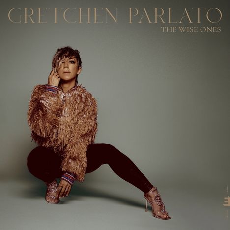 „GRETCHEN PARLATO, THE WISE ONES“. Eine Person sitzt in glamourösem Outfit mit Fransenjacke und High Heels.