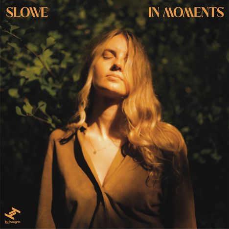 Text: "SLOWE IN MOMENTS". Eine Frau mit geschlossenen Augen steht in der Sonne, umgeben von grünem Laub.
