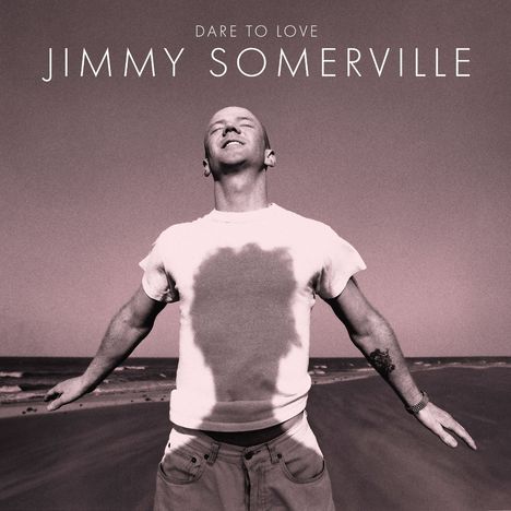 "Dare to Love Jimmy Somerville." Ein Mann mit ausgebreiteten Armen am Strand, trägt ein T-Shirt und blickt lächelnd nach oben.