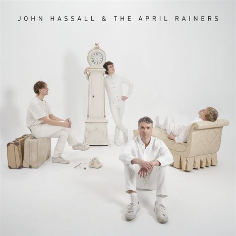 "JOHN HASSALL & THE APRIL RAINERS." Vier Männer in weißer Kleidung, eine alte Standuhr, ein Sofa und Koffer im Raum.