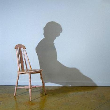 Ein Stuhl wirft einen Schatten, der wie eine sitzende Person aussieht, auf eine helle Wand.