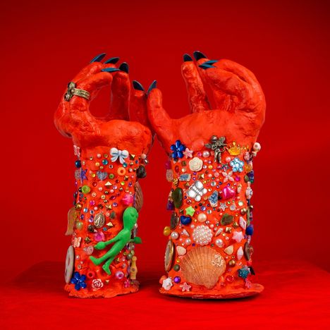 Zwei rote, aufgerichtete Hände, dekoriert mit bunten Perlen, Muscheln und Figuren auf rotem Hintergrund.