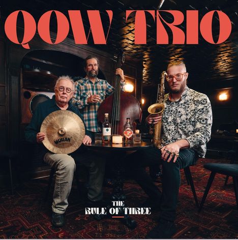 "QOW TRIO" und "THE RULE OF THREE" sind in großen Buchstaben zu sehen. Drei Musiker mit Instrumenten sitzen um einen Tisch.