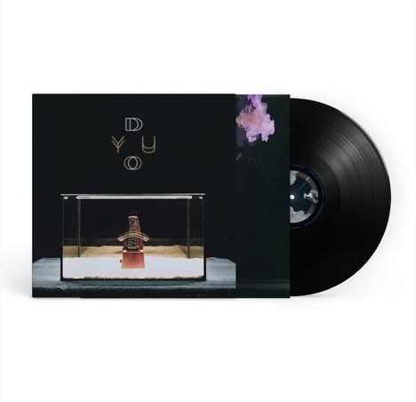 Text: "D Y U O". 
Ein Metronom in einem Glaskasten auf Sand, daneben eine Schallplatte.