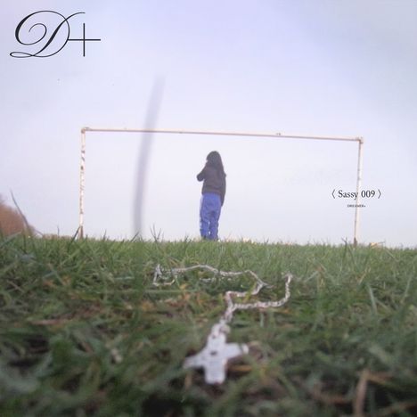 "Sassy 009, Dreamer." Eine Person in blauer Kleidung steht vor einem leeren Tor auf einem Grasfeld. D+ Logo links.