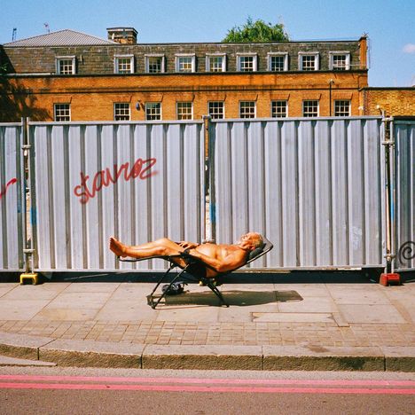 Graffiti mit dem Wort "stavoz", ein Mann entspannt auf einem Liegestuhl vor einer Wellblechwand auf dem Gehweg.