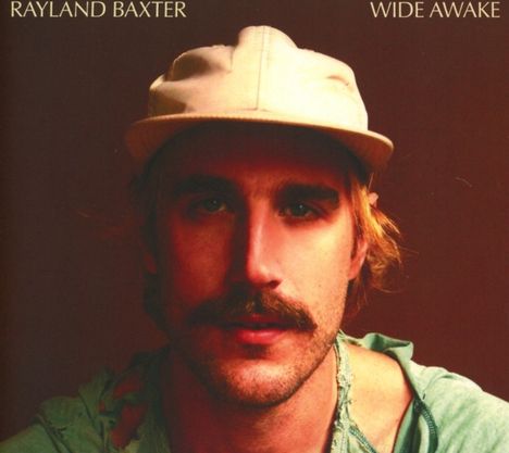 "Rayland Baxter Wide Awake" ist oben geschrieben. Ein Mann mit Schnurrbart und Basecap schaut direkt.