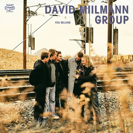 "DAVID MIILMANN GROUP, YOU BELONG." Fünf Menschen stehen lachend nah beieinander vor Bahngleisen in einer ländlichen Umgebung.