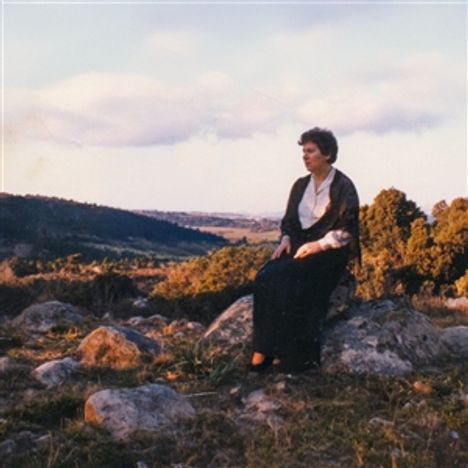 Das Bild zeigt eine Frau, die auf einem Felsen sitzt, umgeben von einer weitläufigen, hügeligen Landschaft bei Sonnenuntergang.