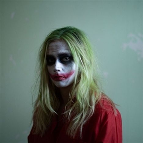 Person mit grünem Haar und Clown-Makeup, das stark betont rote Lippen und schwarze Augen hat. Kleidung in leuchtendem Rot.