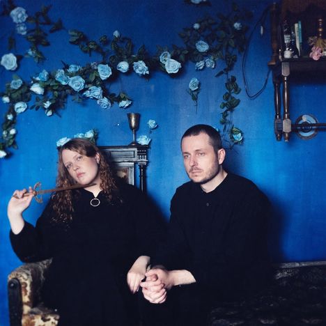 Zwei Personen in schwarzer Kleidung sitzen vor einer blauen Wand mit blauen Rosen, eine hält ein Zepter.