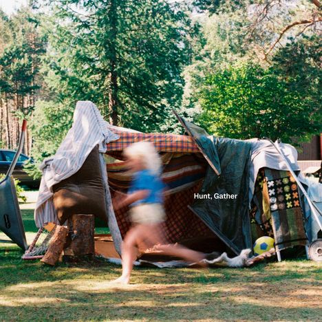 Text: "Hunt, Gather". Eine Kinderspielhütte aus Decken und Stöcken im Garten, ein Kind läuft daran vorbei.