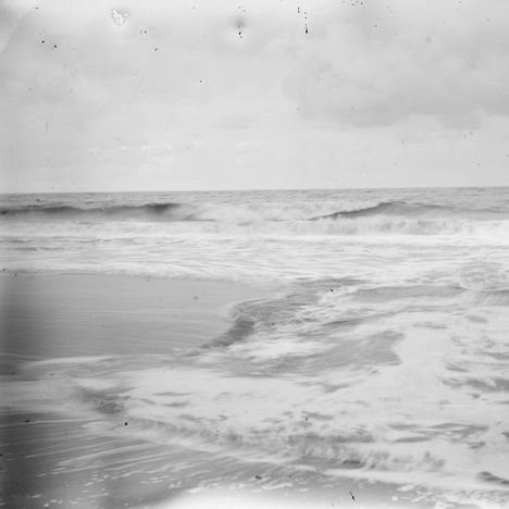 Keine erkennbaren Texte. Wellen brechen sanft an einem Strand unter einem bewölkten Himmel.