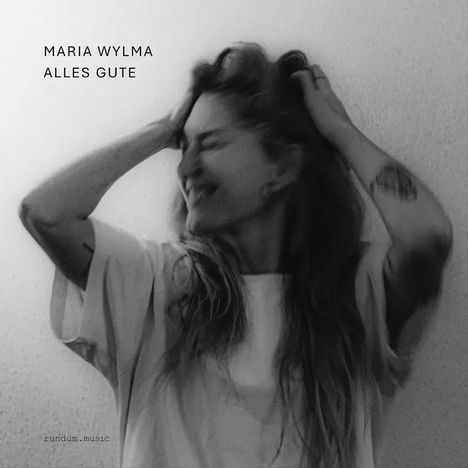 Texte: "MARIA WYLMA ALLES GUTE". Schwarz-weiß Foto einer Frau, die lächelnd die Hände im Haar hat.