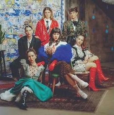 Sechs Personen in extravaganter, bunter Kleidung; manche sitzen, andere stehen. Hintergrund mit Kunstwerken.