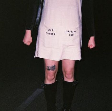 Text auf einem Kleid: „SELF HATRED“ und „MASSIVE EGO“. Jemand steht mit geballten Fäusten, trägt Stiefel und hat ein Bein-Tattoo.