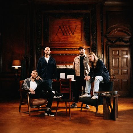 „∆III V“ steht auf Holzplatte, vier Männer in Freizeitkleidung in einem stilvollen Raum mit Holzverkleidung.