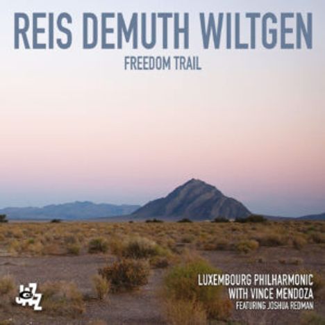 "Reis Demuth Wiltgen", "Freedom Trail". Landschaft mit bergigem Hintergrund und trockenem Gras im Vordergrund. Logo unten links.