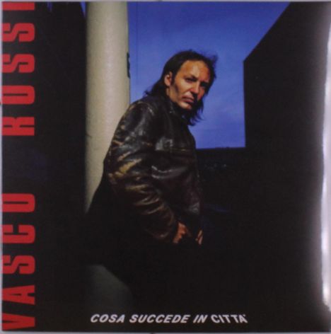 "VASCO ROSSI" steht vertikal, "COSA SUCCEDE IN CITTÀ" unten. Ein Mann in Lederjacke vor urbanem Hintergrund bei Abenddämmerung.