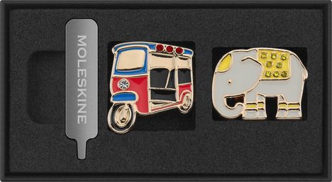 MOLESKINE. Eine silberne Metallklammer, ein bunter Tuk-Tuk-Pin und ein Elefanten-Pin mit gelben Verzierungen.
