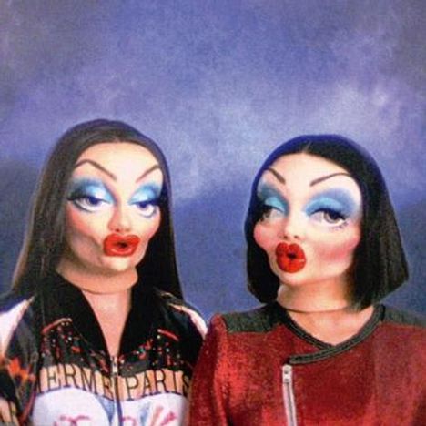 Zwei Figuren mit auffälligem Make-up und puppenartigen Gesichtern, tragen bunte Kleidung.