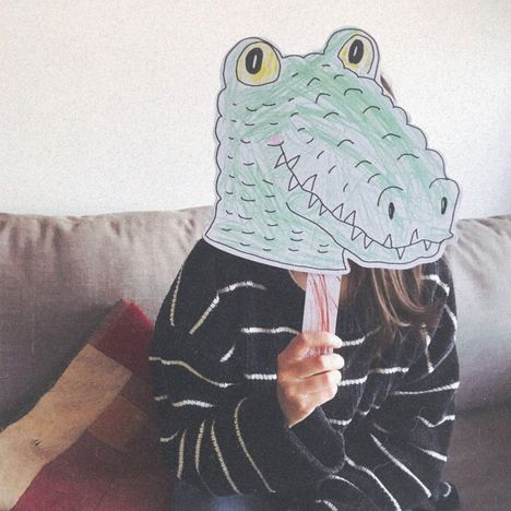 Eine Person hält eine gezeichnete Krokodilmaske vor das Gesicht, sitzend auf einem Sofa.