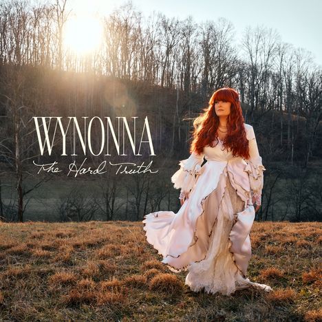 "WYNONNA The Hard Truth." Eine Frau in einem langen, fließenden Kleid mit roten Haaren steht in einer herbstlichen Landschaft.